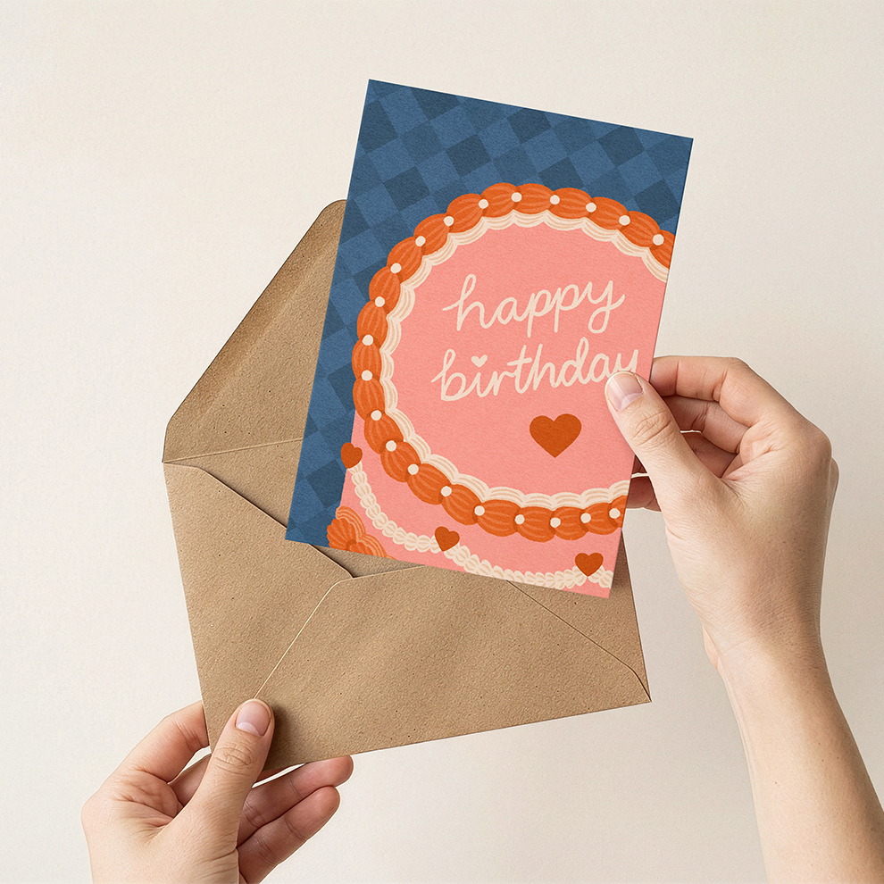 Hand holding a birthday card with a cake design and 'Happy Birthday' text, placed on an open brown envelope.