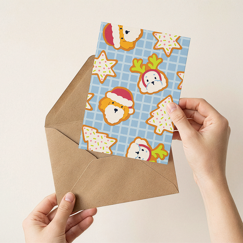 Hand holding a Christmas-themed card with dog, star and tree cookies and an envelope on a beige background