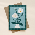 Greeting card with daisy design on a checkered border, placed on a beige surface.