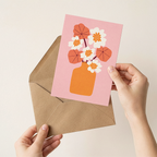 Hand holding a pink card with a floral design and an orange envelope on a beige background