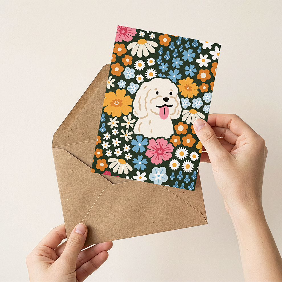 Hand holding a floral card with a dog illustration and a brown envelope on a beige background