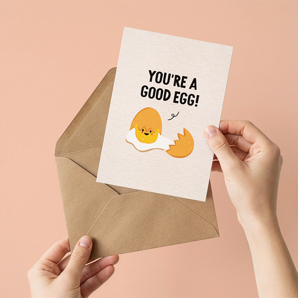 Greeting card with 'You're a good egg!' text and illustration held over a brown envelope on a pink background