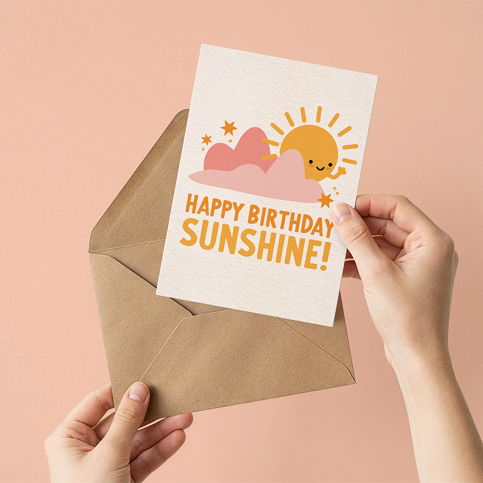 Hand holding a birthday card with a sun and cloud design and 'Happy Birthday Sunshine!' text, against a pink background.