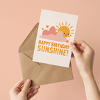 Hand holding a birthday card with a sun and cloud design and 'Happy Birthday Sunshine!' text, against a pink background.