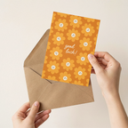 Hand holding a card with 'good luck!' text and floral design, placed inside an open brown envelope on a light background.