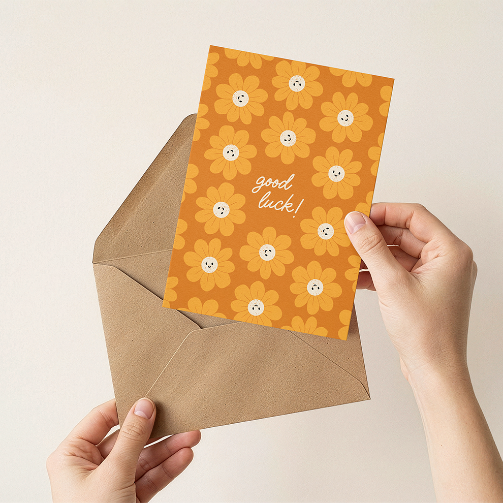 Hand holding a card with 'good luck!' text and floral design, placed inside an open brown envelope on a light background.