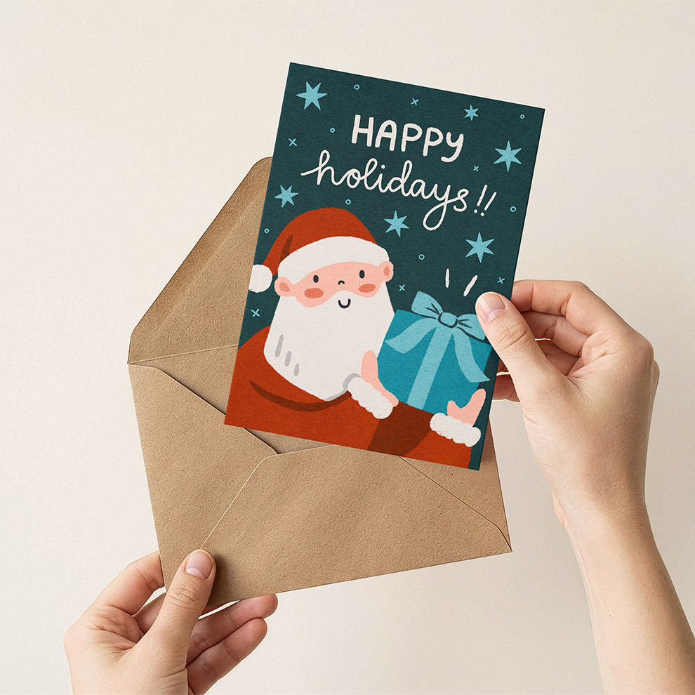 Hand holding a Christmas card with Santa Claus and 'Happy holidays!' text, placed on an open envelope.