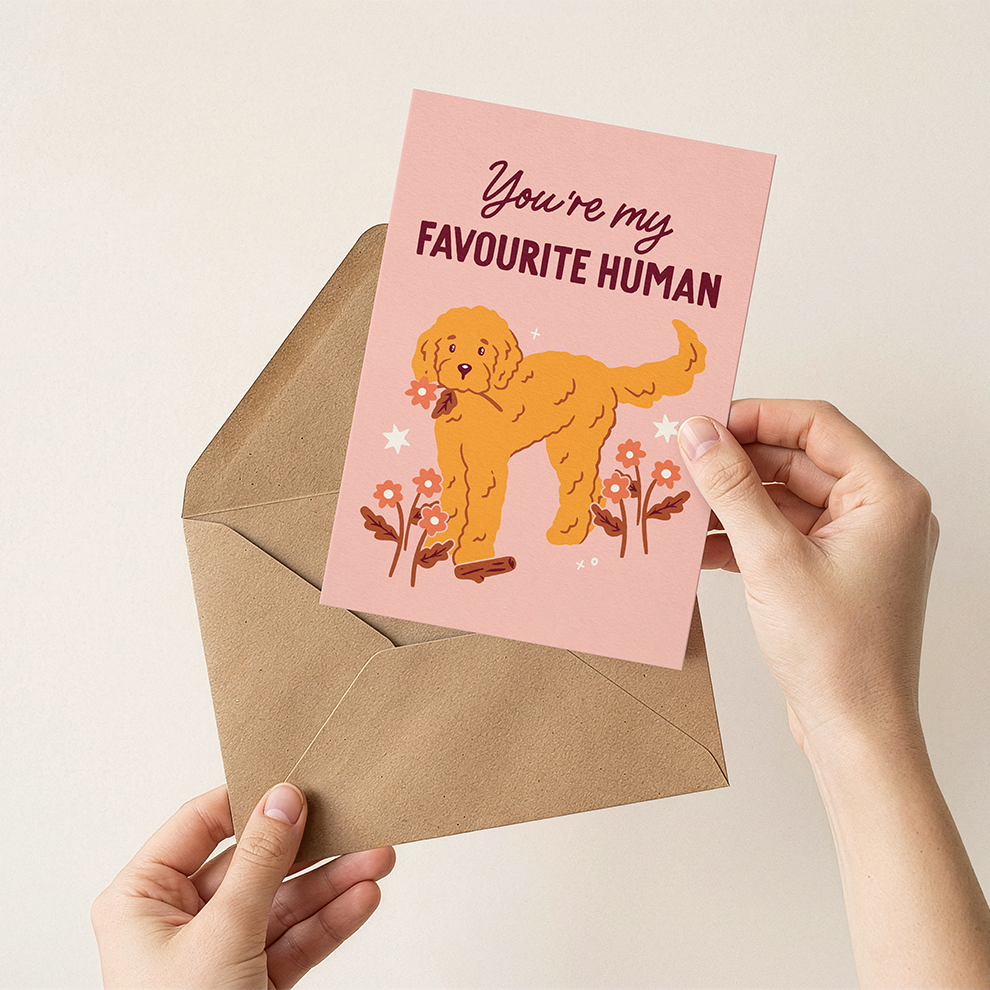 Hand holding a card with a dog illustration and text 'You're my Favourite Human' over a beige envelope.