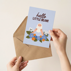 Person holding a greeting card with 'hello little bean!' text and illustrations, next to a brown envelope on a beige background.