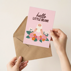 Person holding a pink greeting card with 'hello little bean!' text and an envelope on a beige background