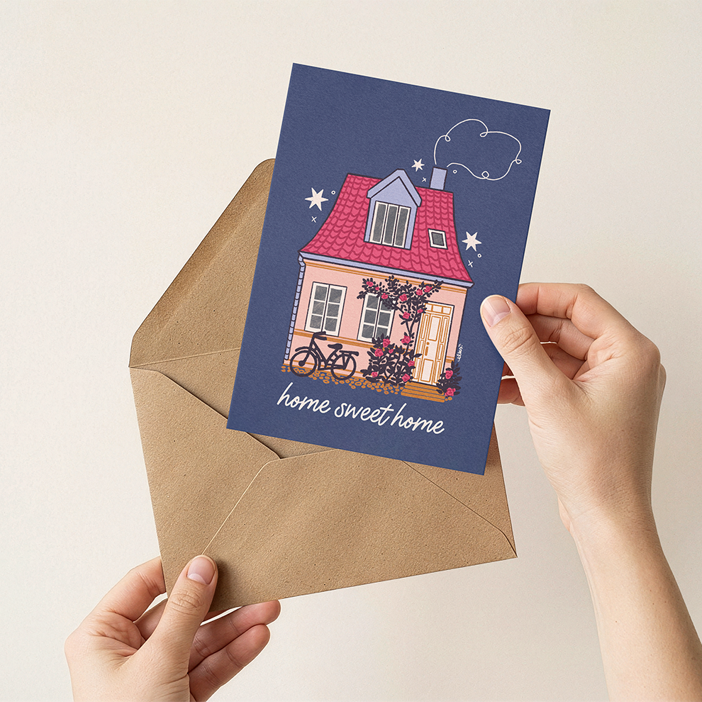 Hand holding a card with a house illustration and 'home sweet home' text, next to an open envelope.