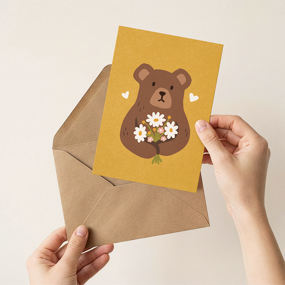 Hand holding a card with a bear illustration and flowers, placed on a brown envelope against a beige background.