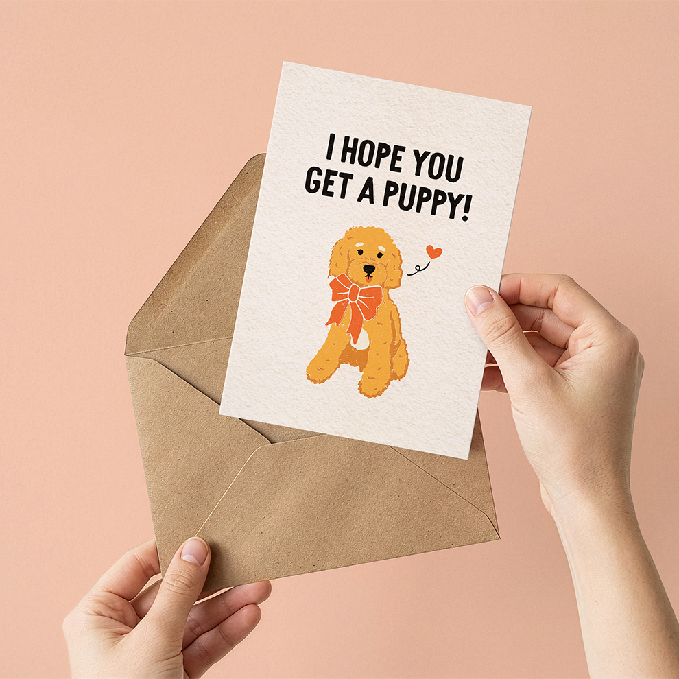 Greeting card with a dog illustration and "I hope you get a puppy" text held over an envelope against a pink background