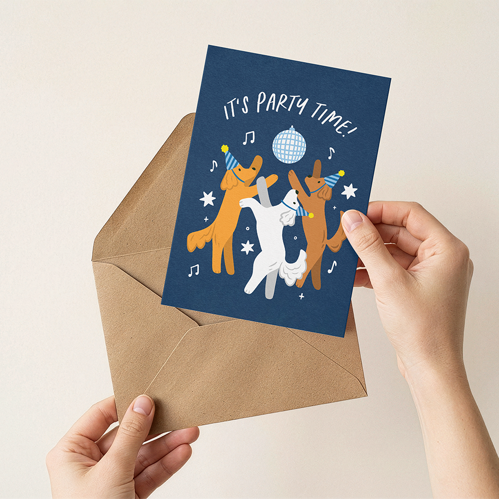 Hand holding a blue card with cartoon dogs and 'It's Party Time!' text, next to a brown envelope on a beige background.