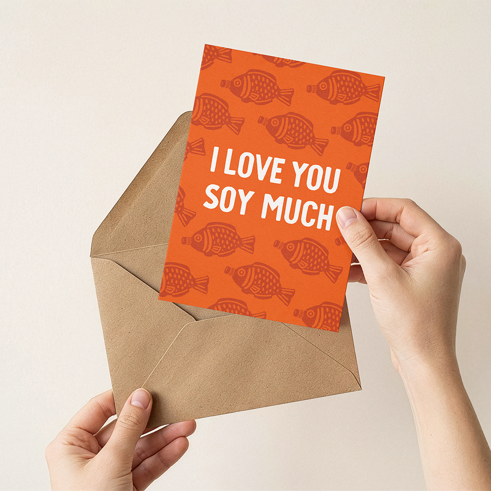 Hand holding an orange card with 'I Love You Soy Much' text and soy sauce fish pattern, placed inside a brown envelope.