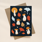 Greeting card with autumn-themed illustrations of mushrooms and leaves on a dark blue background, placed on a beige surface.