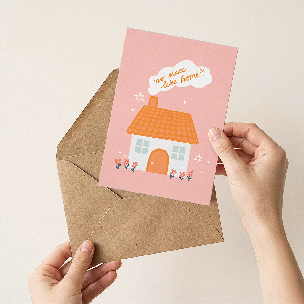 Person holding a pink card with a house illustration and 'No place like home' text, over a brown envelope on a light beige background.