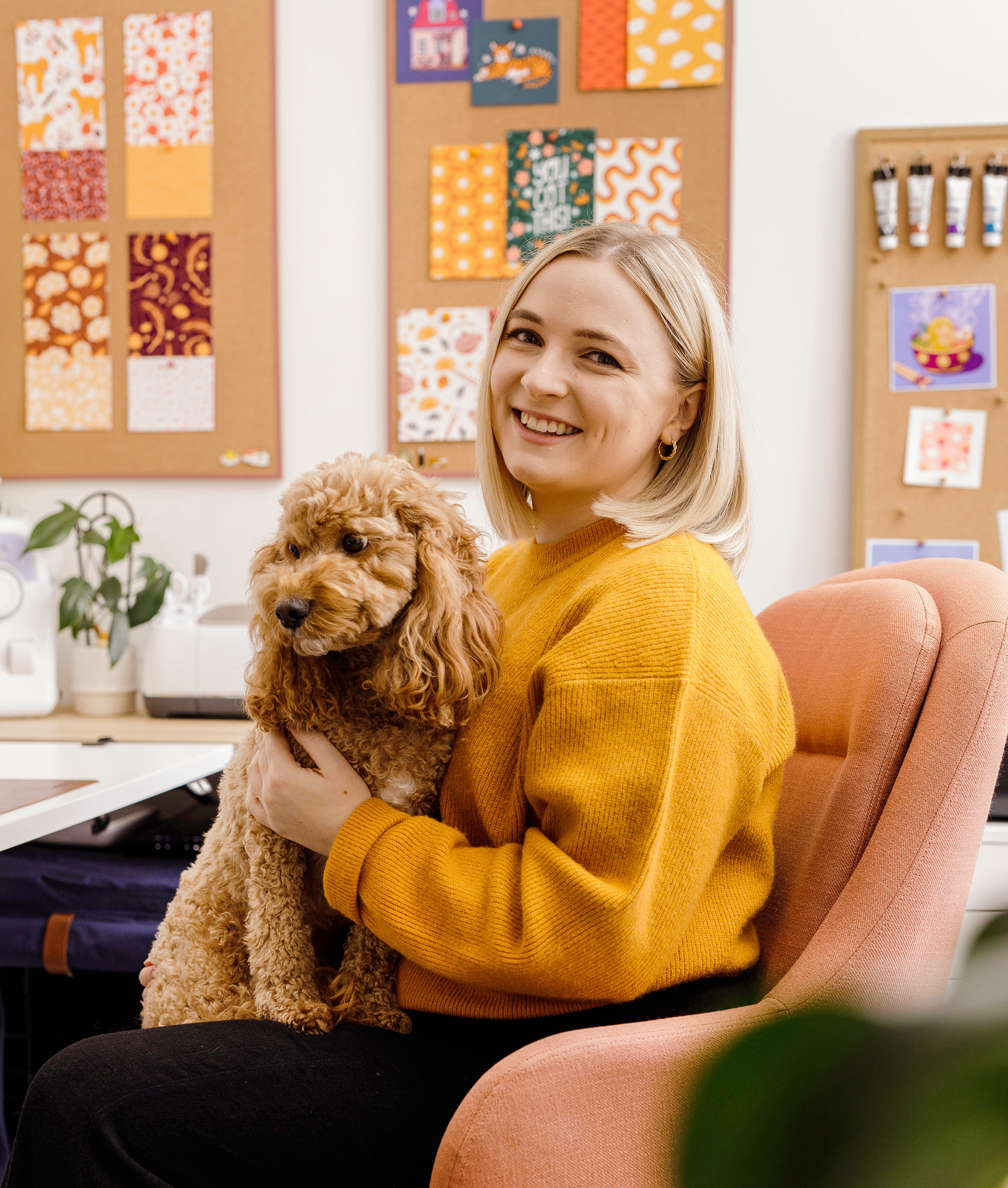Emmi Kerkham from Hello Elks in a yellow sweater holding Winston, a small brown dog, in an indoor setting with colorful art on the wall.