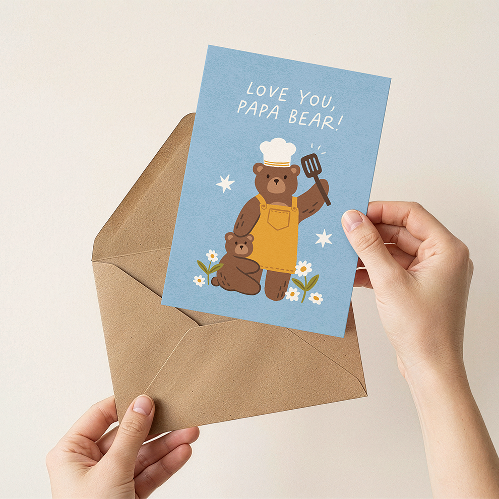 Card with illustration of a bear wearing a chef's hat and apron, holding a spatula, with 'Love you, papa bear!' text, held over a brown envelope.