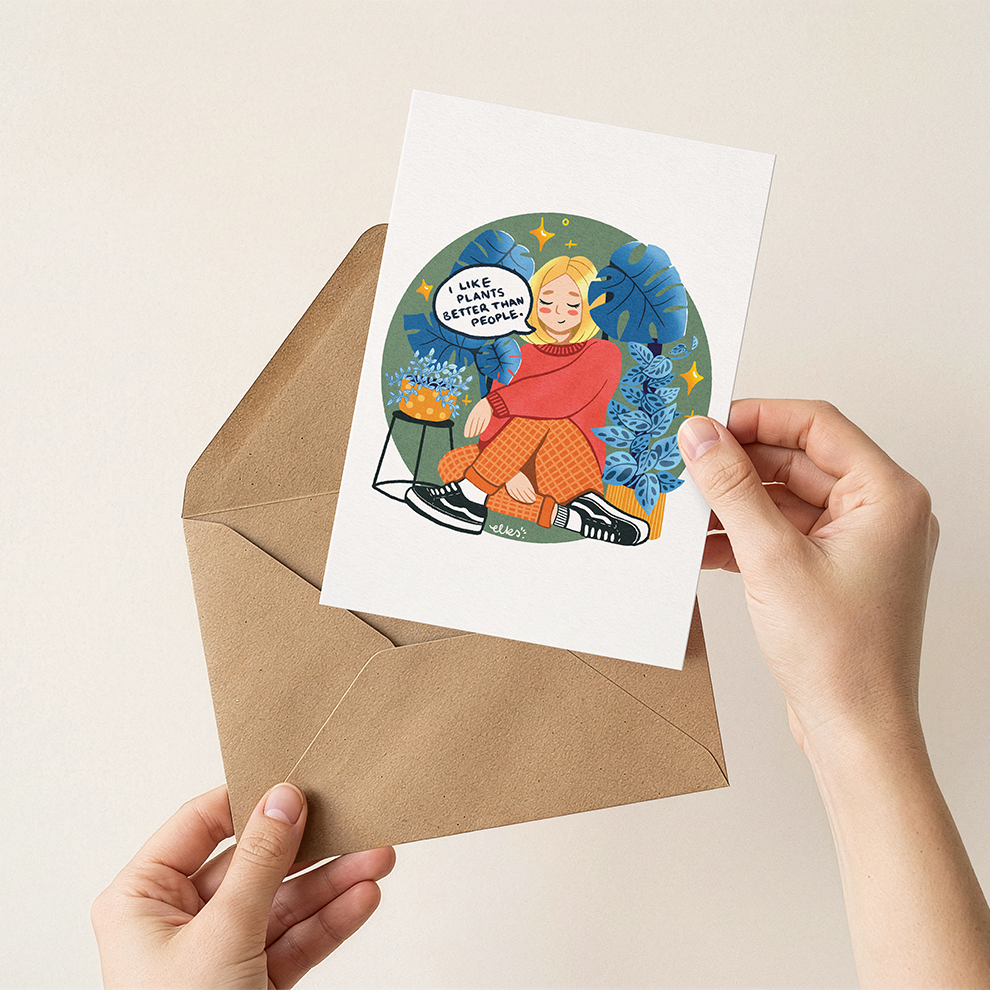 Person holding a greeting card with an illustration of a person in winter clothing, next to a brown envelope on a beige background.