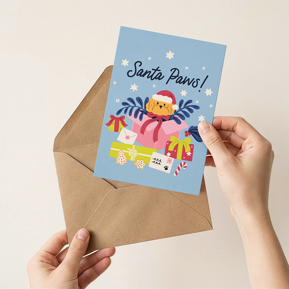Hand holding a Christmas card with a dog and presents, labeled 'Santa Paws', over an envelope.