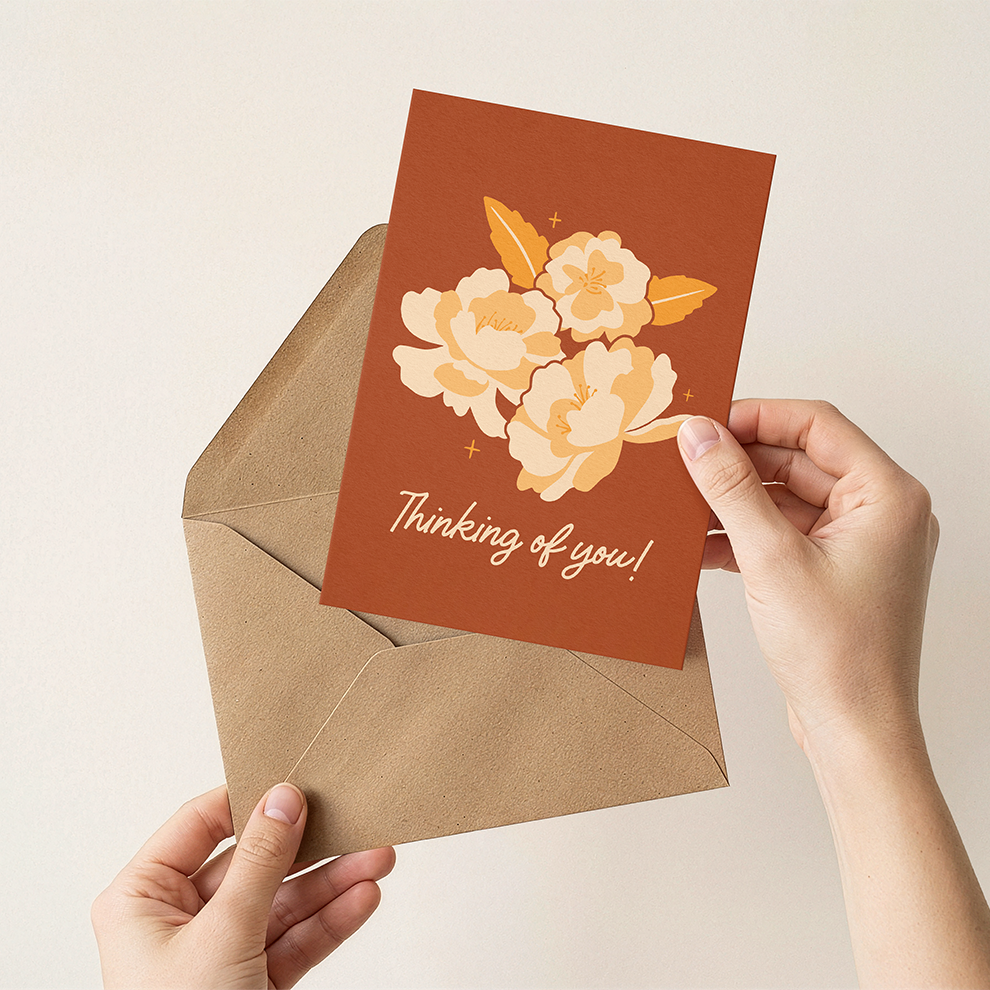 Hand holding a card with floral design and 'Thinking of you!' text, placed on a matching envelope.