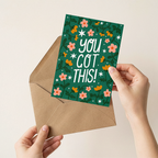 Hand holding a green card with floral designs and 'YOU GOT THIS!' text, placed on a beige envelope.