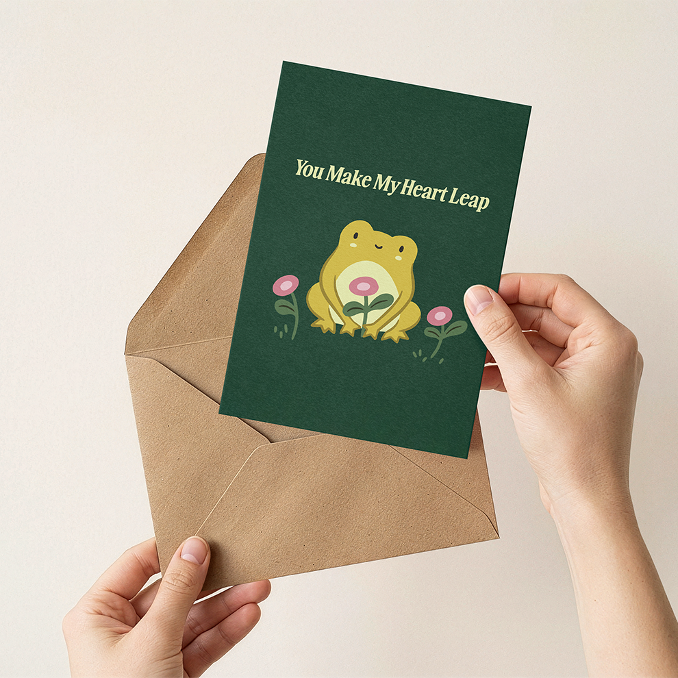 Hand holding a green card with a frog illustration and text, placed on a beige envelope.